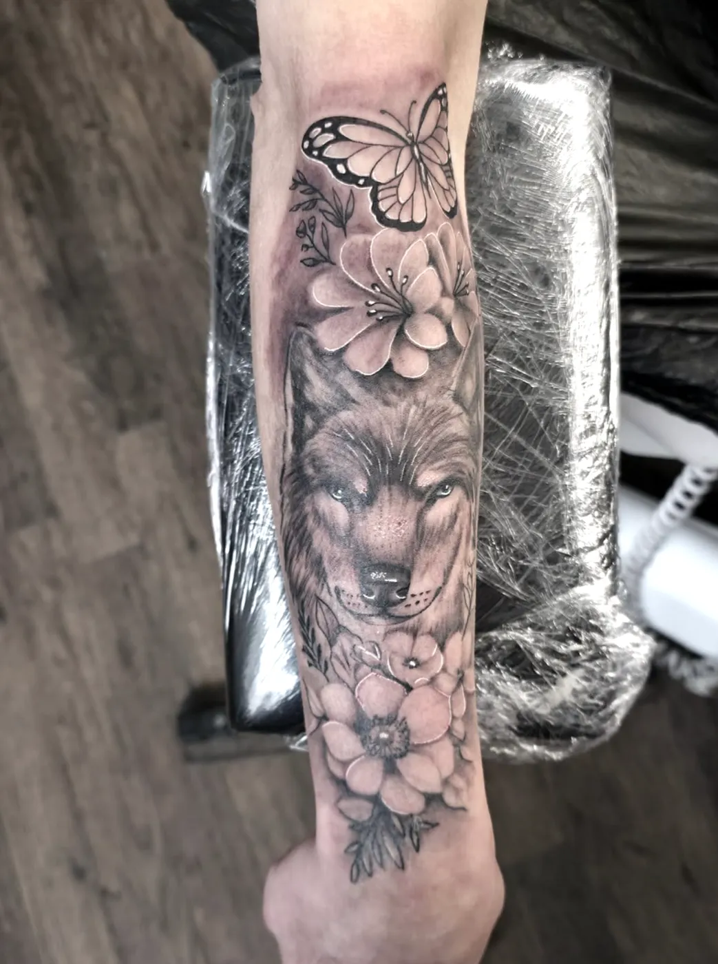 Wolf mit Kirschblüten und Schmetterling als weiches Black-&-Grey-Tattoo auf dem Unterarm.