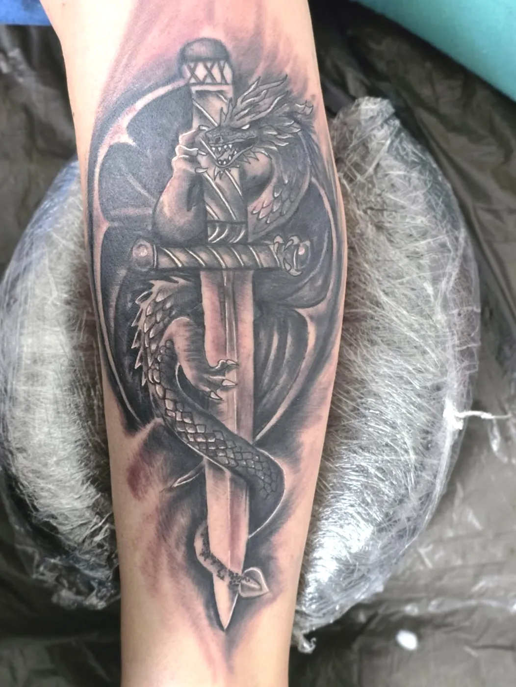 Drache, der sich um einen Dolch windet, als Black-&-Grey-Tattoo auf dem Unterarm.
